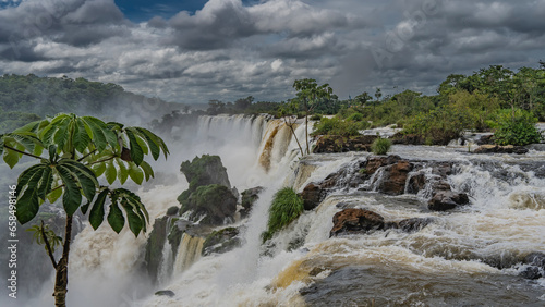 Impressive view of the waterfall. Streams collapse from ledges with splashes. Fog and haze. Picturesque boulders in the riverbed. Tropical vegetation and cloudy sky. Iguazu Falls. Argentina.