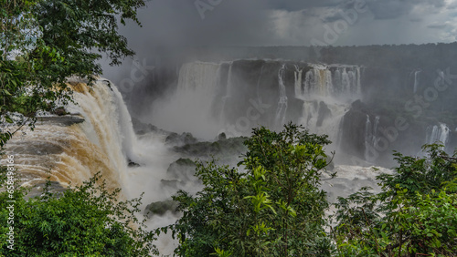 Fototapeta Naklejka Na Ścianę i Meble -  Cascades of waterfalls fall from the ledges of rocks into the river, foaming. Spray and fog rise into the sky. Green vegetation in the foreground. Iguazu Falls. Brazil.