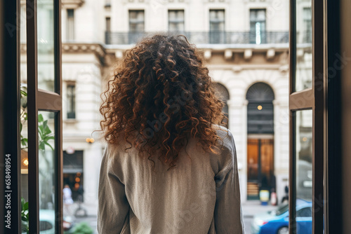 Rear view of a young woman with curly hair looking through the window in the city