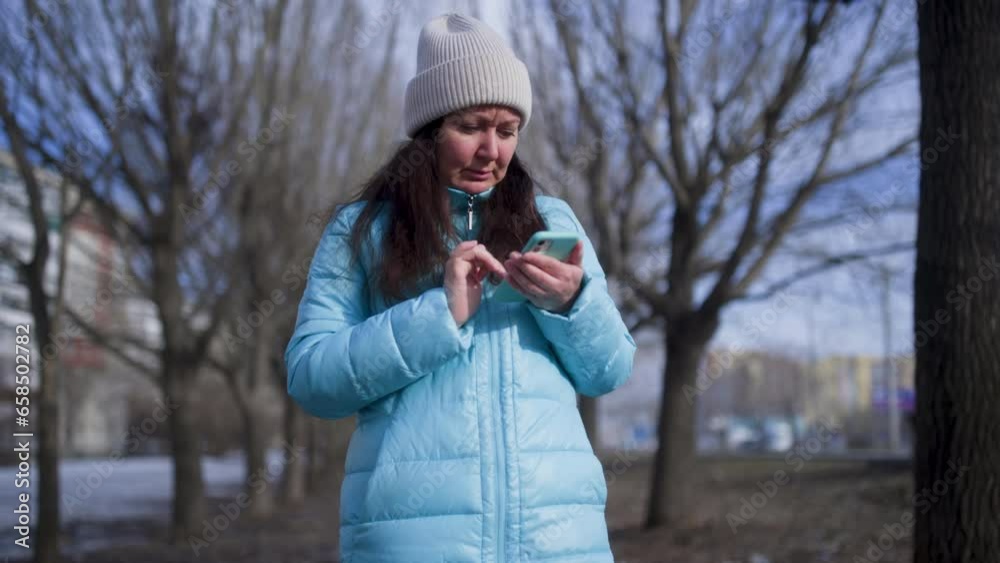 Woman using smartphone while standing against bare trees. Female in warm clothing scrolling internet at park. She is spending leisure time during winter. Woman using smartphone while standing against bare trees. Female in warm clothing scrolling internet at park. She is spending leisure time during winter.