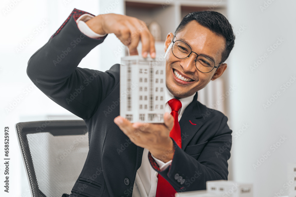 Agent presents contracts for property purchase or lease. Businessman asian indian people showcases miniature model home, signifying secure property insurance. Desk scene in a home sales office. Agent presents contracts for property purchase or lease. Businessman asian indian people showcases miniature model home, signifying secure property insurance. Desk scene in a home sales office.