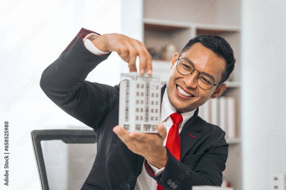 Agent presents contracts for property purchase or lease. Businessman asian indian people showcases miniature model home, signifying secure property insurance. Desk scene in a home sales office. Agent presents contracts for property purchase or lease. Businessman asian indian people showcases miniature model home, signifying secure property insurance. Desk scene in a home sales office.