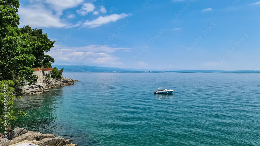 A small boat parked along the coastal line in Lovran, Croatia. Steep rocks going into the sea, overgrown with lush green trees. The high mountains are visible on the horizon. Clear, summer day. Sunny