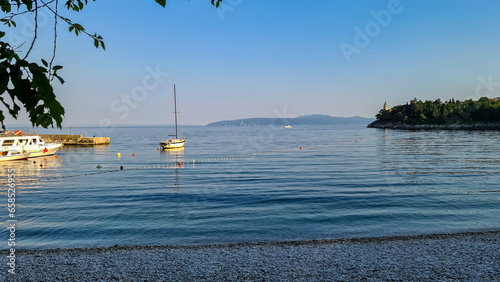 Sailboat parked along the shores of Medveja, Croatia. There are plenty of buoys in the water. The Mediterranean Sea is calm. Stony beach and lush green peninsula on the side. Summer holidays