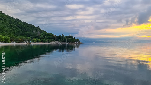 A stony beach along the shore of Medveja in Croatia. The Mediterranean Sea is calm and clear. There is a lush forest with a small town at the shore. The sky is painted yellow. Sunset time. Calmness