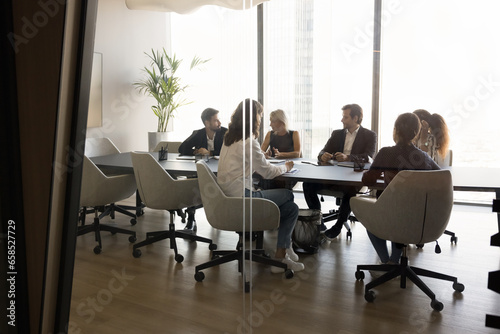 Diverse millennial team of business partners discussing deal in modern office meeting room interior. Group of professionals, entrepreneurs discussing cooperation. Through glass view