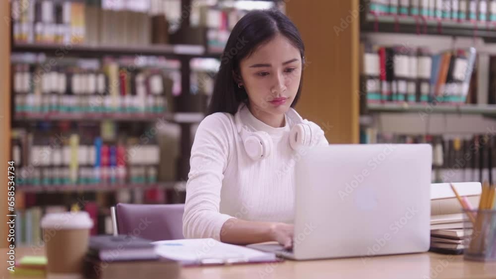 Concentrated Asian teen women student sitting at desk and using laptop for online learning class, and writing notes while preparing for exam in library campus. Concentrated Asian teen women student sitting at desk and using laptop for online learning class, and writing notes while preparing for exam in library campus.