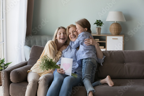 Cheerful happy girls and women of three family generation celebrating grandma birthday, mothers day, 8 march, hugging on home sofa with closed eyes, holding greeting card with heart, flowers