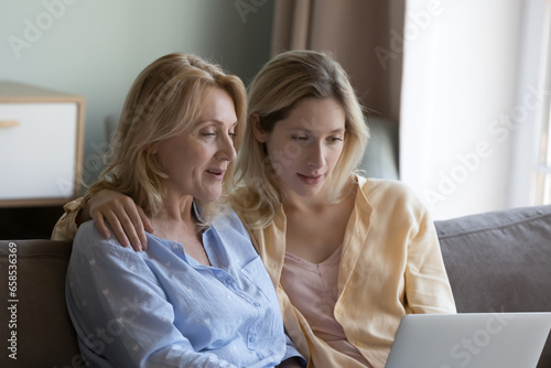 Focused positive mature mother and beautiful young grownup daughter using online ecommerce app on laptop, watching movie, interactive TV channel, enjoying domestic wireless connection, communication