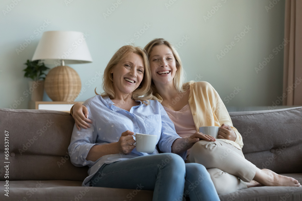 Happy pretty blonde senior mom and young adult daughter drinking hot beverage, coffee, tea at home together, resting on sofa, hugging, looking at camera, smiling, laughing. Family female portrait