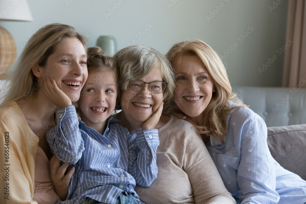 Happy cheerful kid, young, mature and elder women relaxing on sofa, hugging, laughing, looking away. Four female generations sitting, relaxing on couch, enjoying meeting. Home family portrait