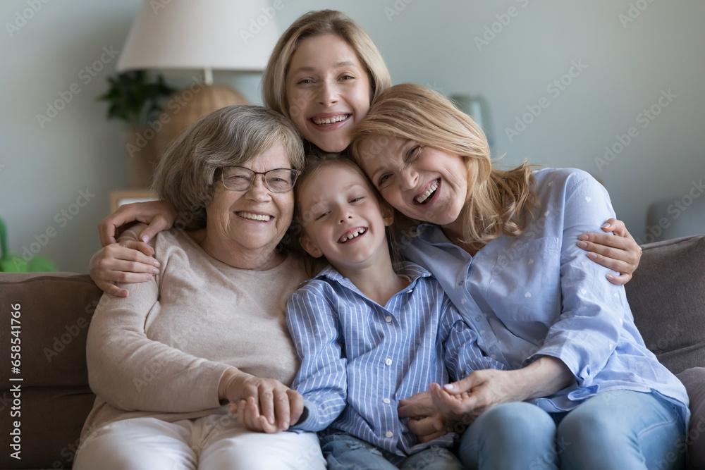 Happy united female relatives of four generations meeting at home, hugging on sofa, looking at camera with toothy smile, laughing, having fun. Girl, mom, granny, great grandmother portrait