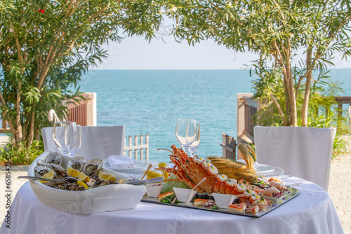 romantic table decorated with flowers for two with glasses and plate with lobster and fresh oysters outdoors in front of the sea on a sunny day.