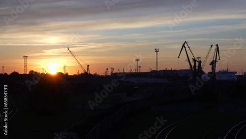 The sun hides behind the silhouettes of harbor warehouses and cranes