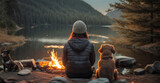 An attractive view of the lake at sunset is being admired by a woman and her dog as they relax next to a campfire. An alliance between a dog and a human