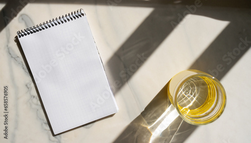 Aesthetic yellow morning flat lay. Glass with water, blank notebook on marble table background. Natural dappled light shadows. Minimal business template, copy space