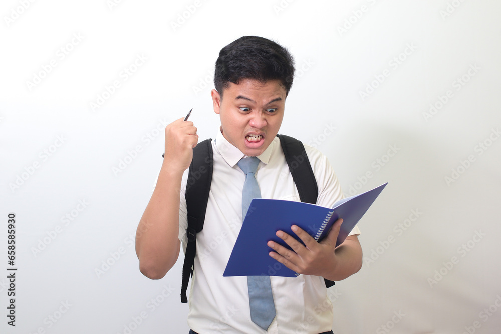 Indonesian senior high school student wearing white shirt uniform with gray tie writing on note book using pen with annoyed and frustrated expression. Isolated image on white background Indonesian senior high school student wearing white shirt uniform with gray tie writing on note book using pen with annoyed and frustrated expression. Isolated image on white background