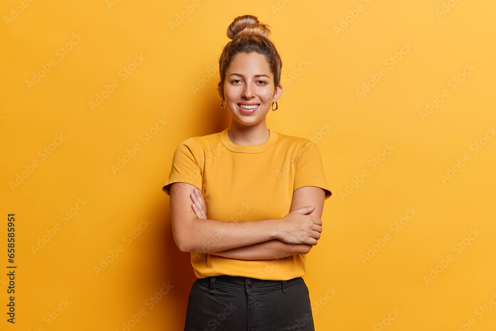 Φωτογραφία Horizontal shot of lovely European woman with hair bun keeps arms folded stands satisfied poses for making photo against vivid yellow background dressed in casual t shirt and black trousers