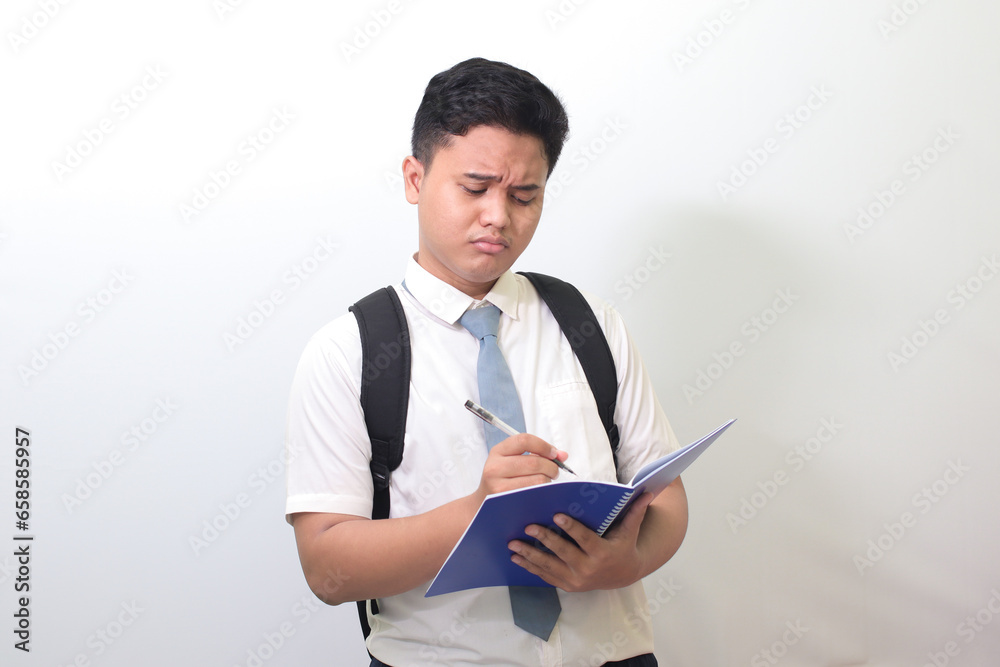 Indonesian senior high school student wearing white shirt uniform with gray tie writing on note book using pen with annoyed and frustrated expression. Isolated image on white background Indonesian senior high school student wearing white shirt uniform with gray tie writing on note book using pen with annoyed and frustrated expression. Isolated image on white background