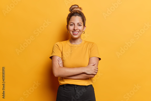 Horizontal shot of lovely European woman with hair bun keeps arms folded stands satisfied poses for making photo against vivid yellow background dressed in casual t shirt and black trousers. photo