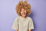 People and sincere emotions concept. Overjoyed curly haired woman has fun and laughs happily looks positively at camera dressed in casual t shirt isolated on purple background hears something positive