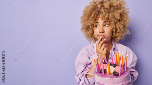 Horizontal shot of beautiful curly haired woman keeps hand on chin concentrated aside poses with tasty cake and burning candles celebrates special occasion isolated over purple background copy space