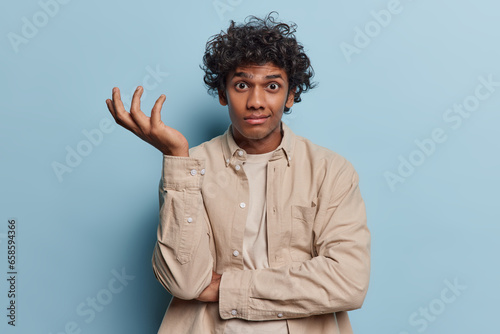 Confused careless man shrugs shoulder with hesitation wears beige shirt says I dont know what to looks clueless poses against blue background. Unsure Hindu guy asks who knows. Difficult choice