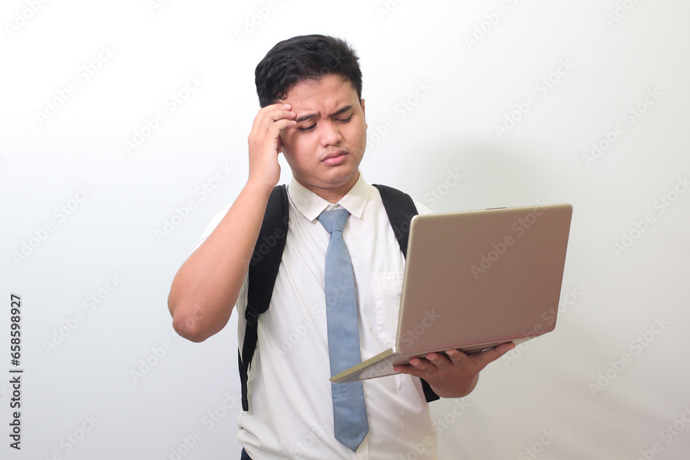 Indonesian senior high school student wearing white shirt uniform with gray tie holding a laptop and showing upset expression, standing against gray background Indonesian senior high school student wearing white shirt uniform with gray tie holding a laptop and showing upset expression, standing against gray background