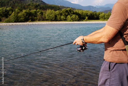 Spinning fishing. Male hands hold a spinning rod against the background of a mountain river, mountains, and blue sky in summer on a sunny day, close-up.