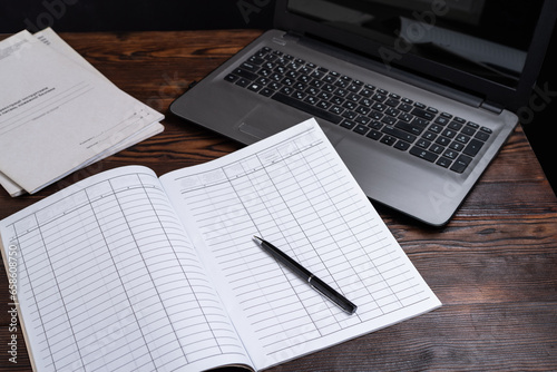 Journal for maintaining financial records. Ballpoint pen and magazine. Notebook and laptop on the table.