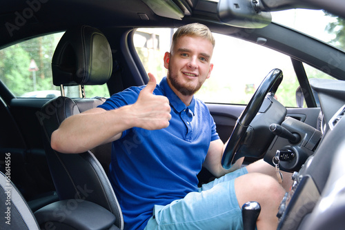 Young happy man sitting in his new car indoors and show thumb up, like gesture. Satisfied customer.