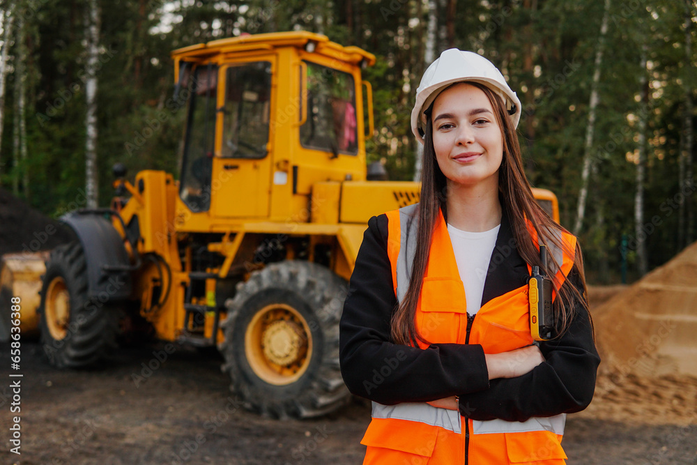 A young woman mine worker is standing in front of a large dump truck or grader. Looking at camera.