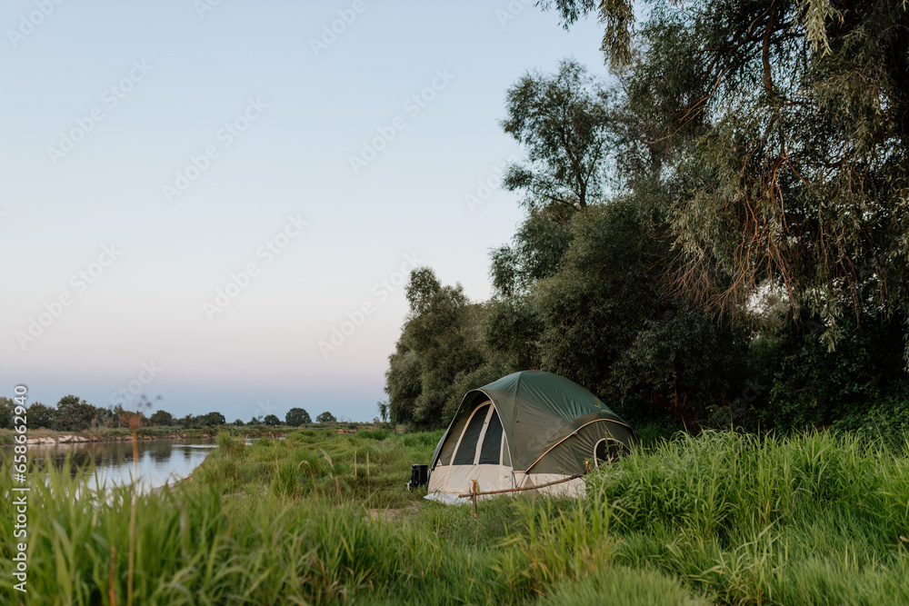 Beautiful summer landscape by the river in the early morning. The tent stands among green grass and trees, on the shore of a lake. Camping. Meeting the dawn by the river.