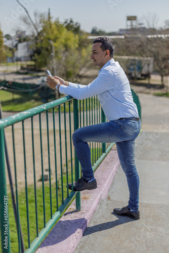 Latino man in a shirt leaning on the railing of a pedestrian bridge using his mobile phone.