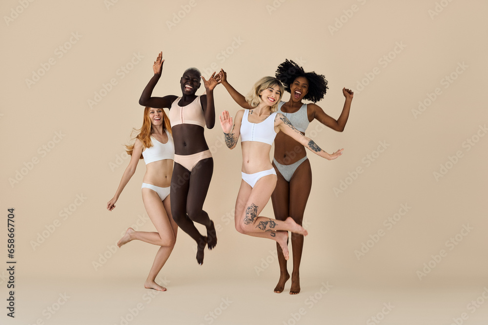 Happy funny diverse girls wearing underwear jumping on beige background. Funky pretty multicultural young women models group laughing having fun, diversity and natural body beauty, authentic shot.