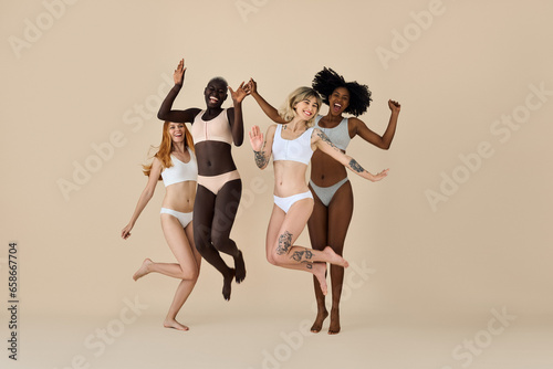 Happy funny diverse girls wearing underwear jumping on beige background. Funky pretty multicultural young women models group laughing having fun, diversity and natural body beauty, authentic shot.