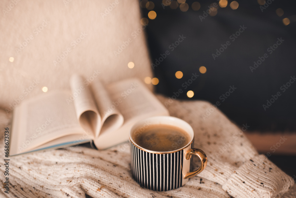 Obraz na płótnie Open paper book with folded pages in heart shape and cup of coffee on knitted clothes over Christmas lights in room close up. Winter cozy home atmosphere. w salonie