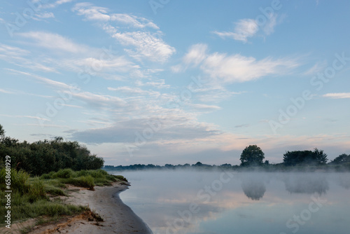 Beautiful summer landscape by the river in the early foggy morning. The tent stands among green grass and trees, on the shore of a lake. Camping. Watching dawn near a river covered in thick fog
