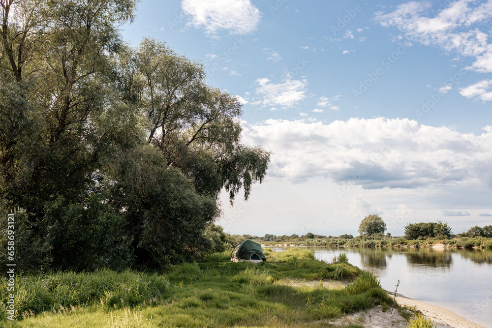 Obraz premium Beautiful summer landscape by the river on a sunny summer day. The tent stands among green grass and trees, on the shore of a lake. Camping.