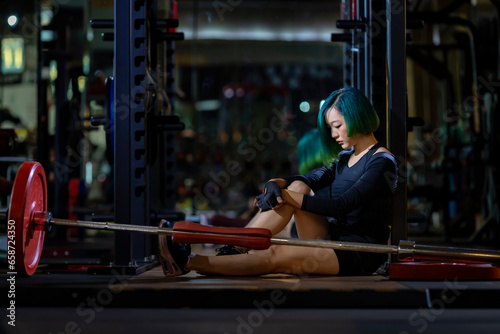 Asian woman is facing overtraining syndrome after weight training workout inside gym with dark background for exercising and fitness concept