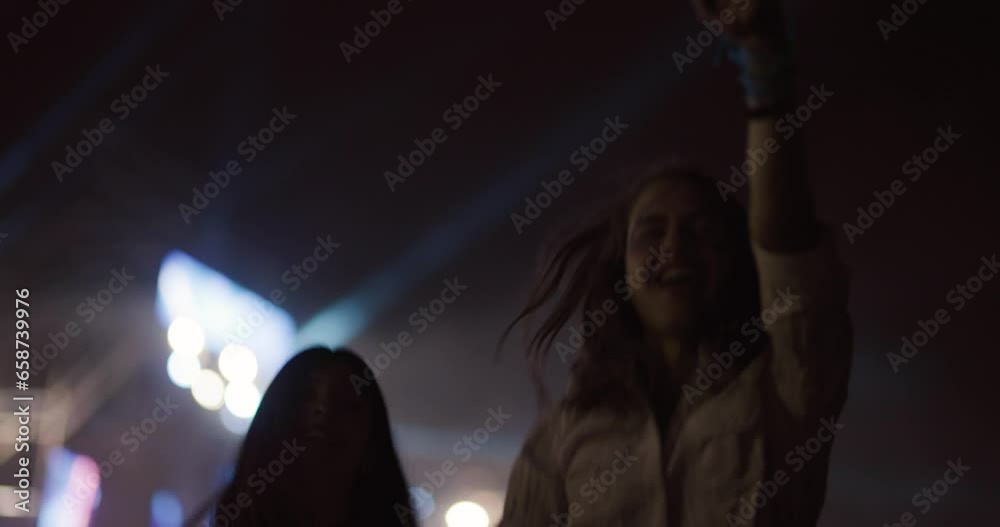 Two female friends dancing together on open space party with flashing lights. Happy beautiful girls jumping with led strip lights on arms. Out of focus crowd and big stage in the background.