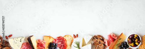 Assortment of charcuterie cheeses, meats and appetizers. Top view bottom border on a white marble background with copy space.