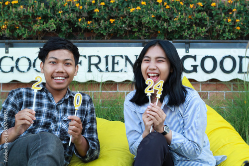happy asian couple teen holding number 20 24 golden candle and sitting on yellow bean bag in house yard, look to camera photo