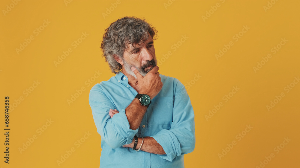 Elderly gray-haired bearded man wears a blue shirt, makes a difficult decision, isolated on an orange background in the studio Elderly gray-haired bearded man wears a blue shirt, makes a difficult decision, isolated on an orange background in the studio