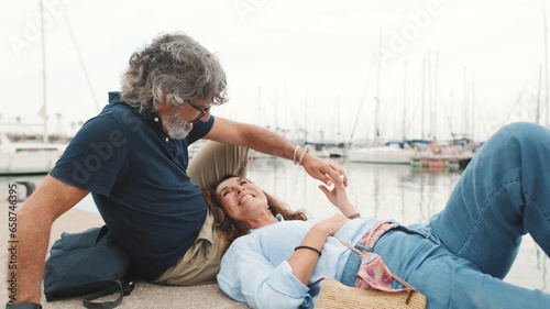 Laughing middle aged couple, retired elderly couple enjoy outdoor recreation together, relax and chat while lying on the pier