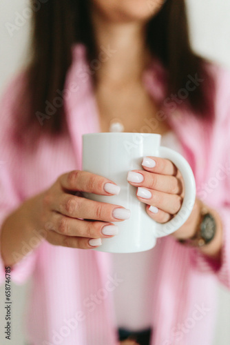 A young girl with black long hair in a pink shirt and white top holds white mug in a cozy room. A white mug in the hands of a girl. Morning business woman