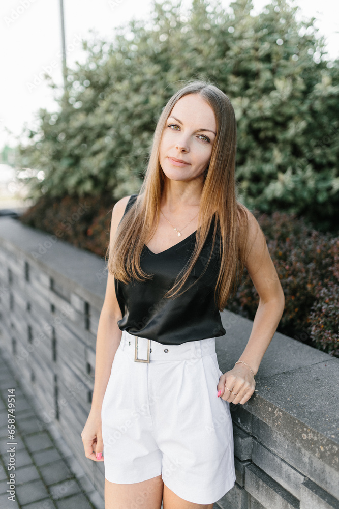 Portrait of a girl with long blond hair, wearing a black top, white shorts, poses against a gray wall. Young woman walking through a park with modern architecture and landscape design Portrait of a girl with long blond hair, wearing a black top, white shorts, poses against a gray wall. Young woman walking through a park with modern architecture and landscape design