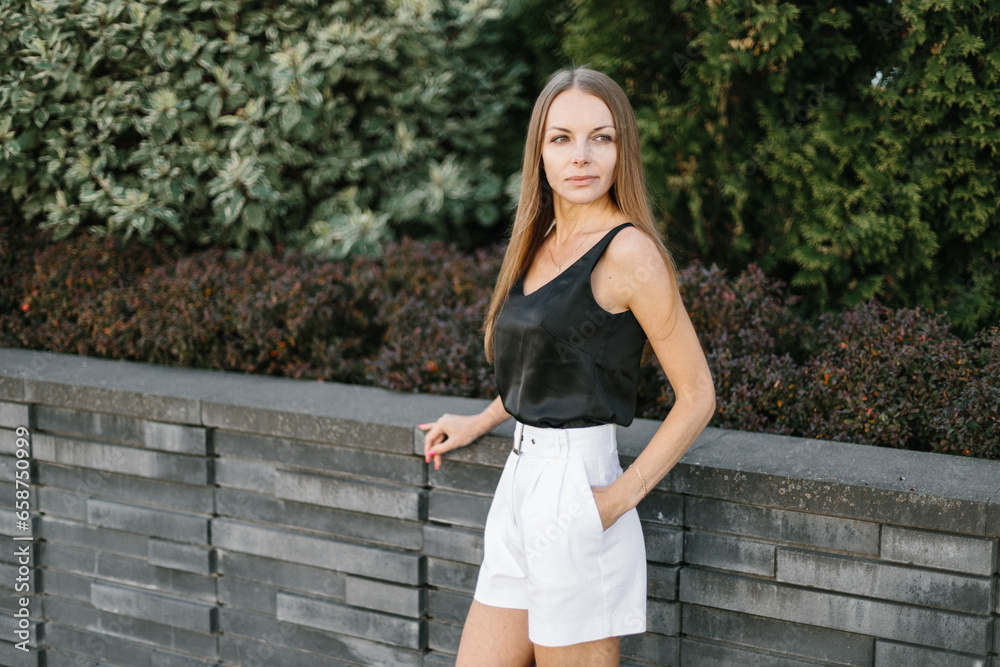 Portrait of a girl with long blond hair, wearing a black top, white shorts, poses against a gray wall. Young woman walking through a park with modern architecture and landscape design Portrait of a girl with long blond hair, wearing a black top, white shorts, poses against a gray wall. Young woman walking through a park with modern architecture and landscape design