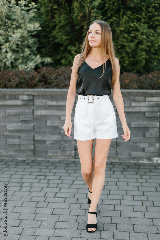 Portrait of a girl with long blond hair, wearing a black top, white shorts, poses against a gray wall. Young woman walking through a park with modern architecture and landscape design