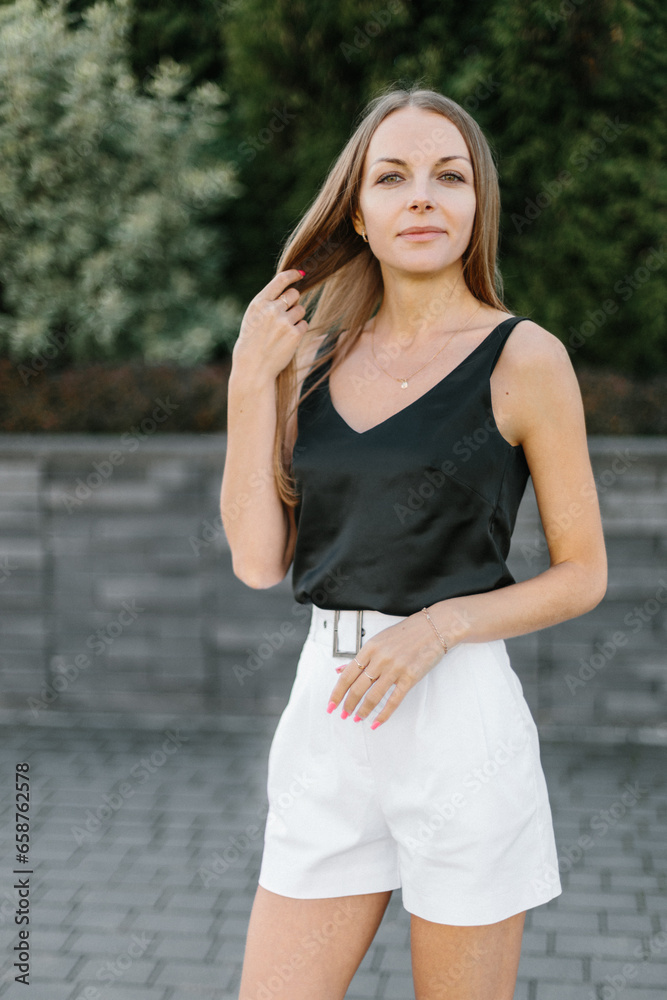 Portrait of a girl with long blond hair, wearing a black top, white shorts, poses against a gray wall. Young woman walking through a park with modern architecture and landscape design Portrait of a girl with long blond hair, wearing a black top, white shorts, poses against a gray wall. Young woman walking through a park with modern architecture and landscape design